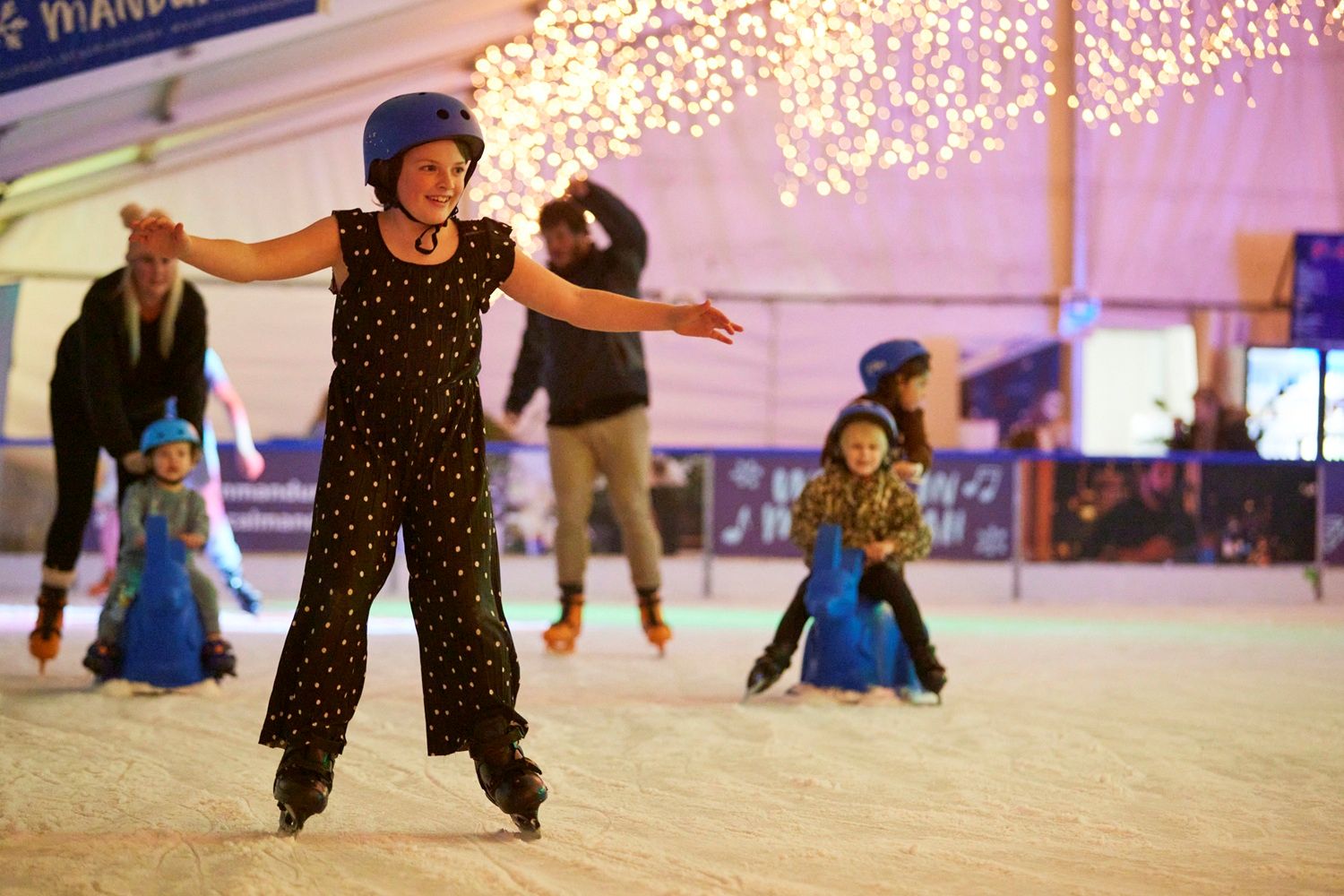 A young girl wearing a blue helmet and a black-and-white polka dot jumpsuit smiles as she ice skates independently at a rink, with other skaters and festive string lights blurred in the background.